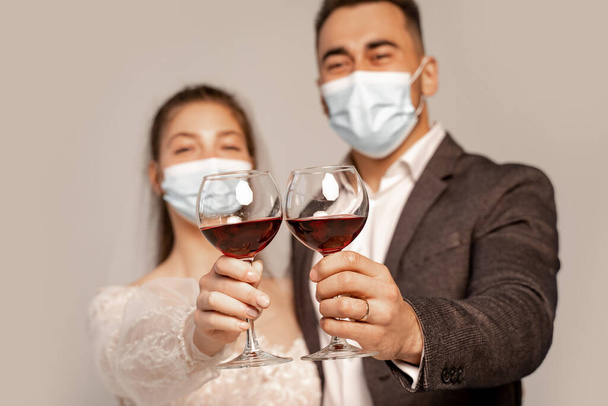 selective focus of glasses with red wine in hands of newlyweds in medical masks isolated on grey - Photo, Image