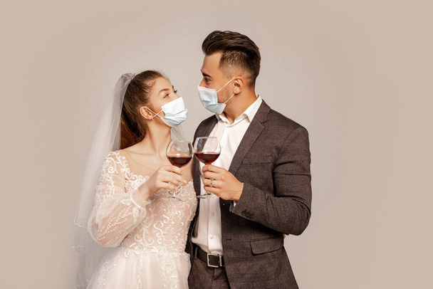 newlyweds in safety masks looking at each other while clinking wine glasses isolated on grey - Foto, Imagem