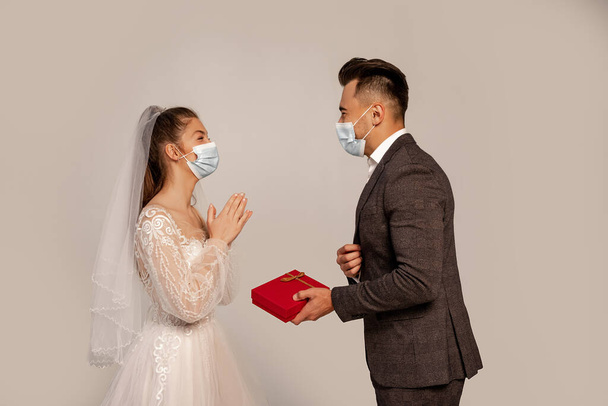 side view of man in medical mask presenting gift box to elegant bride isolated on grey - Photo, Image