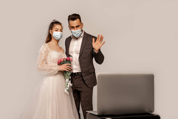 groom in medical mask waving hand during video call on blurred laptop isolated on grey - Φωτογραφία, εικόνα