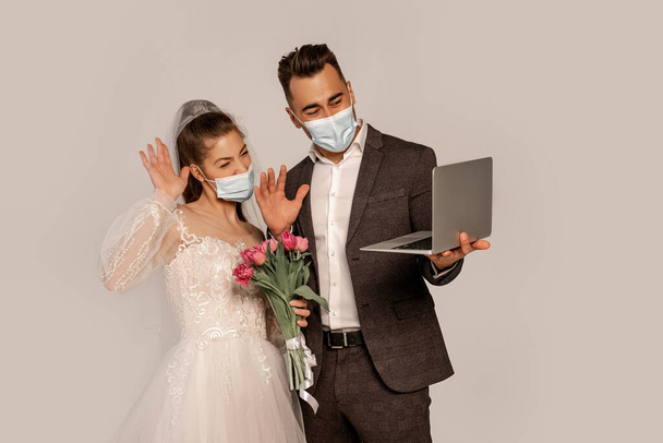 Braut und Bräutigam in medizinischen Masken winken bei Videoanruf isoliert auf grau - Foto, Bild