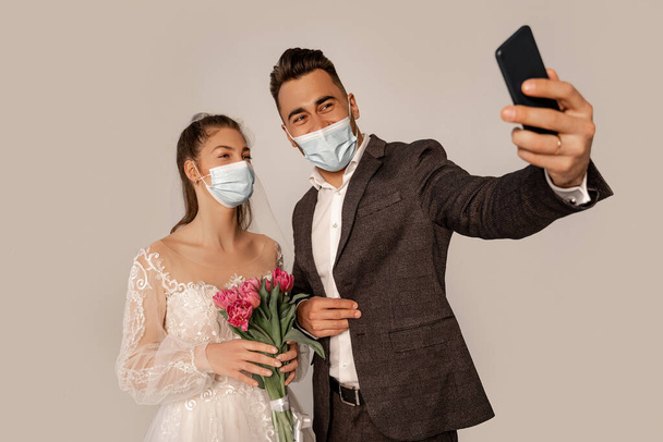newlyweds in safety masks taking selfie isolated on grey with lilac shade - Zdjęcie, obraz
