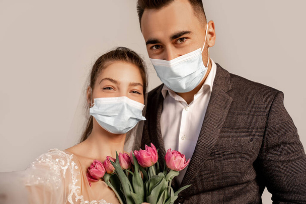 young newlyweds in medical masks looking at camera near fresh tulips isolated on grey - Foto, Imagen