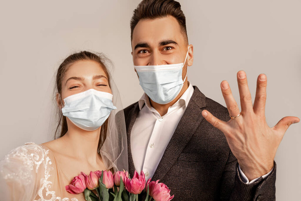 groom in medical mask showing wedding ring near bride with tulips isolated on grey - Foto, Imagen