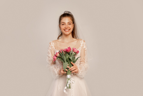 fröhliche Braut mit Tulpenstrauß blickt vereinzelt in die Kamera auf grau - Foto, Bild