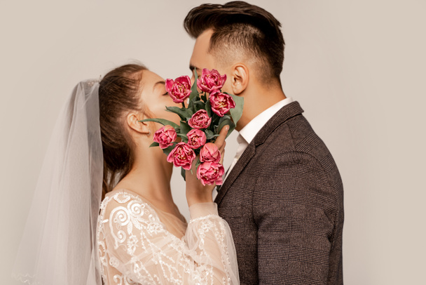 young newlyweds kissing while obscuring faces with tulips isolated on grey - Foto, afbeelding