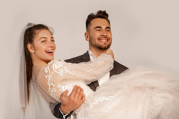 glücklicher Mann hält fröhliche Braut in Händen isoliert auf grau - Foto, Bild