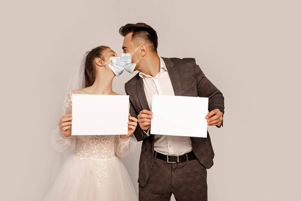 newlyweds in safety masks holding blank cards while kissing isolated on grey - Fotoğraf, Görsel