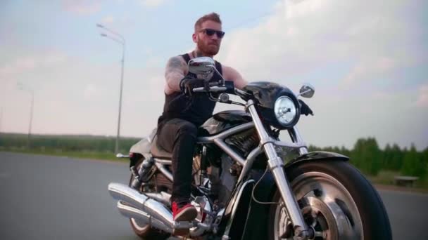 Motociclista elegante con tatuaggi guida una moto su una strada di campagna al tramonto - Filmati, video