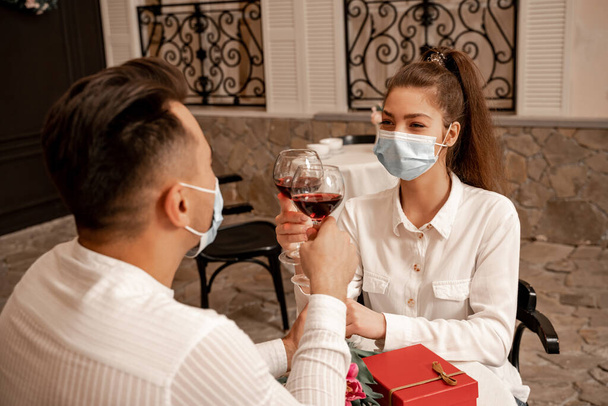 young couple in medical masks clinking wine glasses during date in restaurant - Photo, image