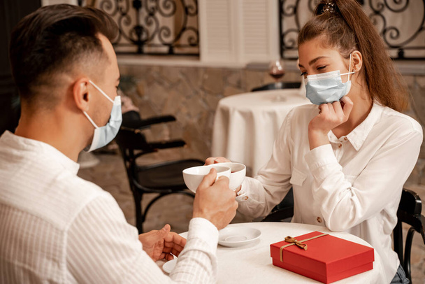 young couple in medical masks clinking cups of tea near gift box on table in cafe - Foto, Bild
