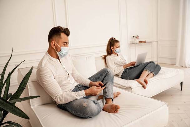 couple in medical masks and jeans using gadgets on couch at home - Foto, Imagen
