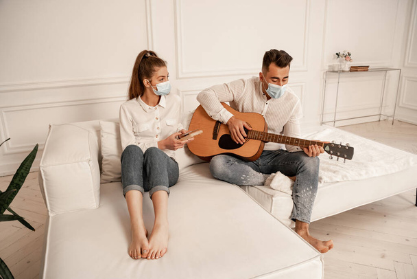пара в медицинских масках и повседневной одежде, играющая на мбире и акустической гитаре дома - Фото, изображение