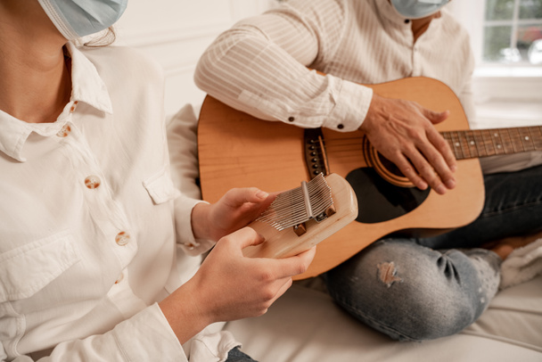Ausgeschnittene Ansicht eines Paares in medizinischen Masken, das zu Hause Musikinstrumente spielt - Foto, Bild
