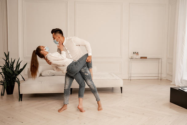 young couple in safety masks and jeans dancing in living room - Foto, imagen