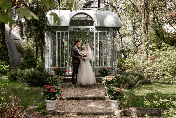 full length view of newlyweds in medical masks standing near alcove in park - Foto, immagini