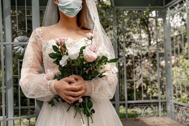 cropped view of elegant bride in medical mask holding wedding bouquet in park - Fotó, kép