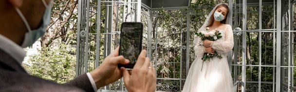 blurred man taking photo of bride in medical mask on smartphone in park, banner - 写真・画像