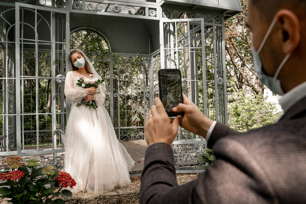 blurred man in medical mask taking picture of bride near alcove in park - Foto, afbeelding