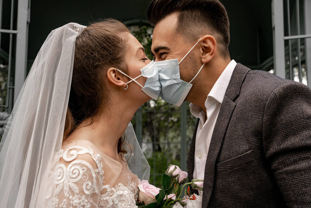 side view of elegant newlyweds in medical masks kissing in park - Фото, изображение
