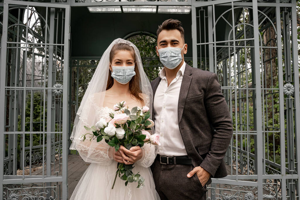 elegant newlyweds in medical masks looking at camera near alcove in park - Φωτογραφία, εικόνα
