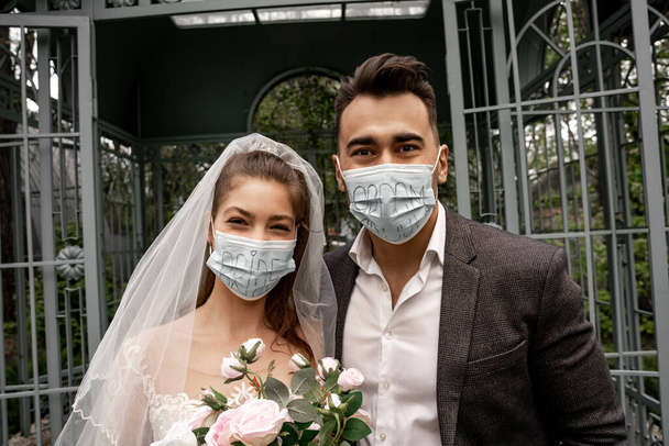 Frischvermählte in medizinischen Masken mit Braut- und Bräutigam-Schriftzug schauen in die Kamera - Foto, Bild