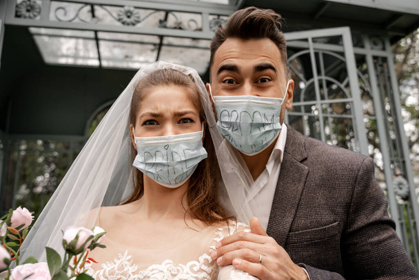 excited newlyweds in safety masks with bride and groom lettering looking at camera in park - Φωτογραφία, εικόνα