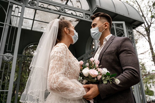 low angle view of newlyweds in protective masks looking at each other outdoors - Foto, Imagem