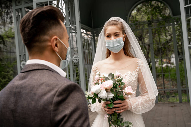 young bride in medical mask holding wedding bouquet near groom on blurred foreground - Foto, Imagem