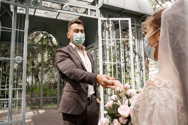 young newlyweds in safety masks holding hands in park on blurred foreground - 写真・画像