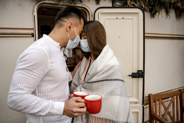 young couple in medical masks standing with cups of coffee and closed eyes near camper - Foto, Imagen