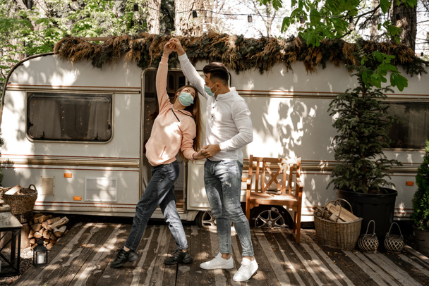 young couple in protective masks dancing near camper in forest - 写真・画像