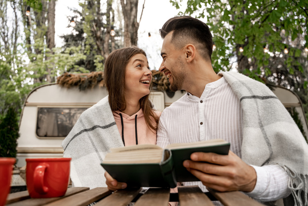 cheerful couple looking at each other while sitting with book in camping - Foto, Bild