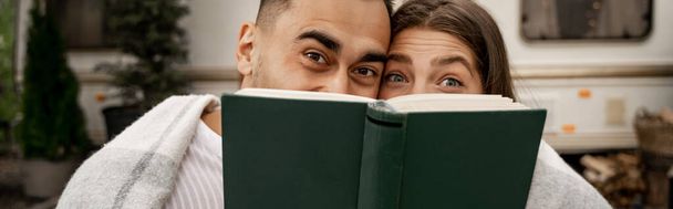 joyful couple obscure faces with book while looking at camera outdoors, banner - Photo, image