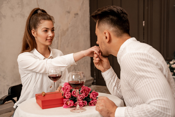 Mann küsst Hand einer hübschen Frau in der Nähe von Weingläsern, Geschenk und Strauß im Restaurant - Foto, Bild