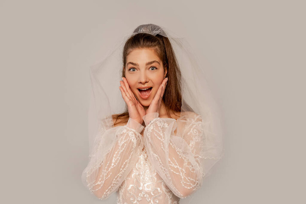 Verblüffte Braut berührt Gesicht und blickt in Kamera isoliert auf grau  - Foto, Bild