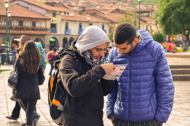 Δύο νεαροί άνδρες τουρίστες που κρατούν και ελέγχουν ένα χάρτη στην πόλη ταξιδιωτικού προορισμού. Κεντρική πλατεία της πόλης (πλατεία στρατώνα) του Κούσκο στο Περού. Φίλοι με τα πόδια περιοδεία το κρύο πρωί - Φωτογραφία, εικόνα