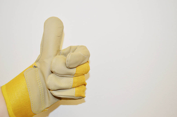 Χέρι σε μπεζ και κίτρινο γάντι ύφασμα εργασίας δείχνει την κατηγορία σημάδι κατά λευκό φόντο στον τοίχο - Φωτογραφία, εικόνα