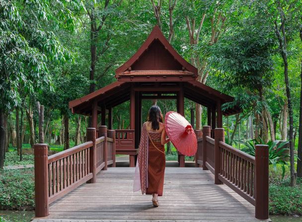 parte posterior de la mujer asiática en vestido tradicional tailandés sosteniendo paraguas rojo caminando en el puente en el antiguo pabellón de madera en el parque del árbol verde - Foto, imagen