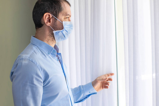 Young man in isolation at home near window. Man in quarantine looking through the window. Covid-19 virus protection. - Foto, imagen