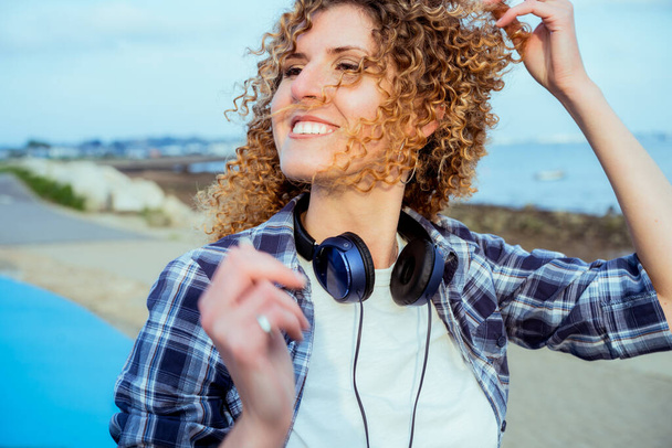 Close up Portrait of smiling curly hair caucasian woman with large headphones walking outdoor. Enjoy the moment and music. Freedom and happiness, simple pleasures concept. - Foto, Imagen