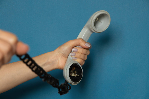 weibliche Hand hält einen alten kaputten Telefonhörer auf einem Draht vor blauem Hintergrund, der Telefonhörer am Prododi ist kaputt - Foto, Bild