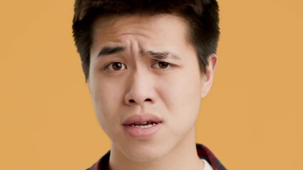 Verwirrter asiatischer Kerl runzelt die Stirn beim Anblick einer Kamera über orangefarbenem Hintergrund - Filmmaterial, Video