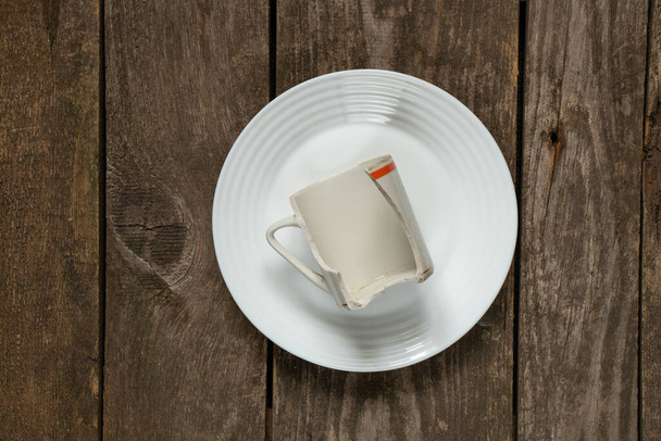 una taza rota yace en un plato blanco sobre una vieja mesa de madera, platos rotos - Foto, Imagen