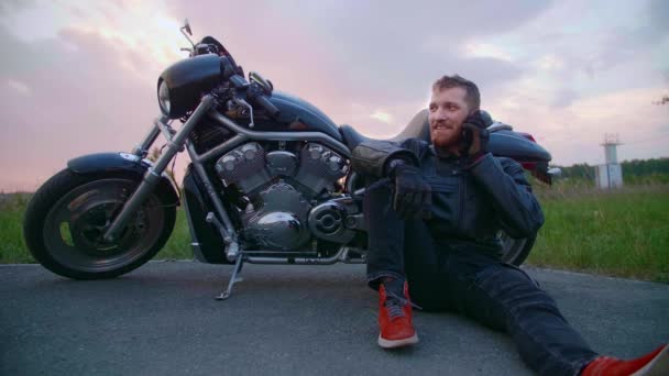 Un motard d'âge moyen assis à côté d'une moto et parle sur son téléphone portable - Séquence, vidéo