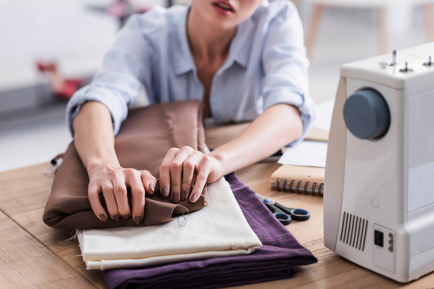 Cropped view of seamstress taking fabric near blurred sewing machine and notebook  - Valokuva, kuva
