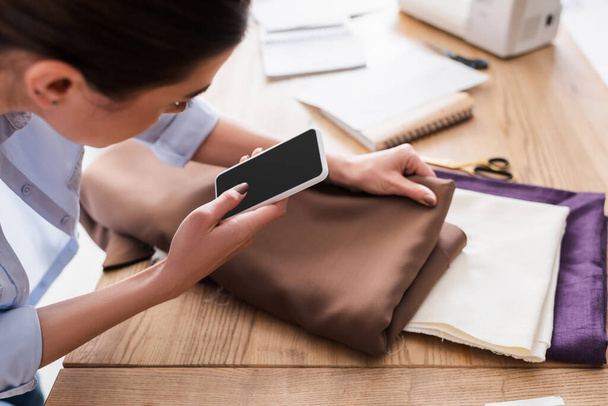 Costurera borrosa con teléfono celular sosteniendo tela cerca de tijeras en la mesa  - Foto, imagen