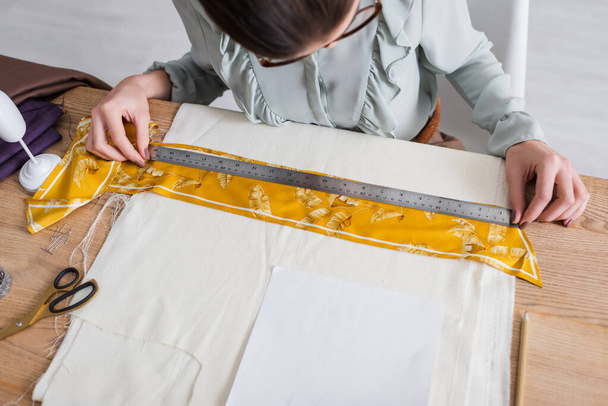 Vista de alto ângulo do designer trabalhando com régua e tecido perto de tesoura na mesa  - Foto, Imagem