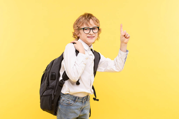 Studio portrait of emotional schoolboy with backpack, isolated on yellow background. Happy child with glasses points a finger to an empty spot. Advertising concept great idea, eyesight, back to school - Photo, Image
