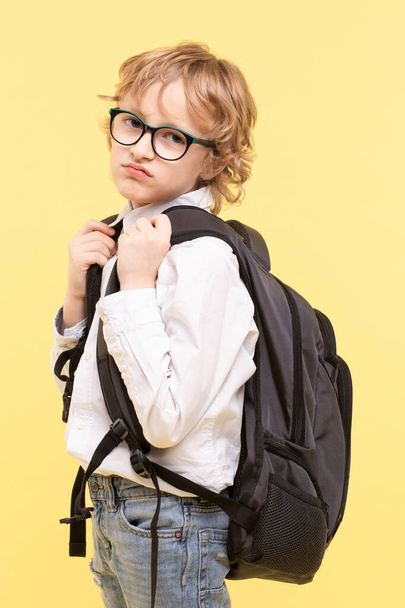 Elementary schoolboy with glasses, large heavy backpack. Boring facial expression. Caucasian boy, blonde on yellow background. Concept  of back to school, problems with school, unwillingness to learn. - Foto, afbeelding